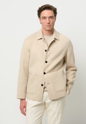 Beige wool jacket with a collar, front pockets, six black buttons, and a straight cut. Worn over a light-colored top and trousers.