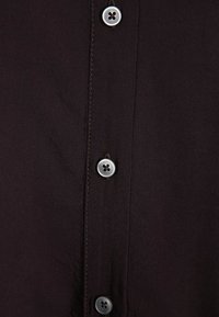 Chemise boutonnée marron foncé avec une texture lisse. Présente des coutures contrastées et des boutons ronds gris à quatre trous. Gros plan sur le tissu.