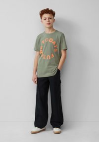 Groene katoenen t-shirt met oranje grafische tekst "DE TOEKOMST VANDAAG" in een cirkelontwerp. Draagt bij zwarte losse cargobroeken en witte sneakers.