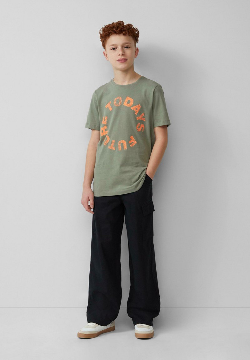 Groene katoenen t-shirt met oranje grafische tekst "DE TOEKOMST VANDAAG" in een cirkelontwerp. Draagt bij zwarte losse cargobroeken en witte sneakers.