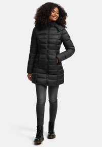 Doudoune noire avec un col haut, design matelassé, fermeture éclair devant et poches. Portée sur un pantalon gris et des bottes noires.