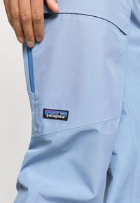 Ανοιχτό μπλε παντελόνι Patagonia με λεία υφή, διαθέτοντας τσέπη με φερμουάρ και μαύρο λογότυπο με πολύχρωμα γραφικά βουνών.