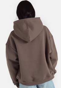 Brązowa, oversize'owa bluza z kapturem na sznurki, z prążkowanymi mankietami i dołem. Gładka faktura, luźny krój, noszona na jasnoniebieskie dżinsy.