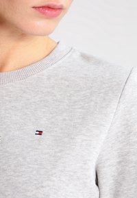 Sudadera gris de algodón con cuello acanalado y un pequeño logo en rojo, blanco y azul en el lado izquierdo del pecho. Textura suave.