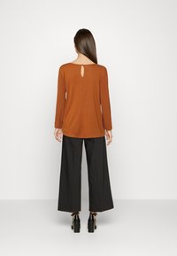 Top marrone a maniche lunghe con dettaglio a chiave sul retro, abbinato a pantaloni neri a gamba larga; tessuto morbido con una vestibilità rilassata.