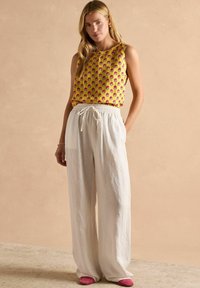 Kollane ilma varrukatop floristiikaga; valged linased püksid elastse vöökohtaga; pehmed roosad slip-on kingad. Neutraalne taust.