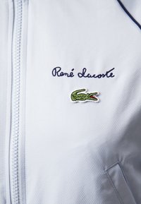 Chaqueta de cremallera azul claro con tejido TEXTURIZADO, que presenta un bordado con la firma 'René Lacoste' y un logotipo de cocodrilo verde en el pecho.