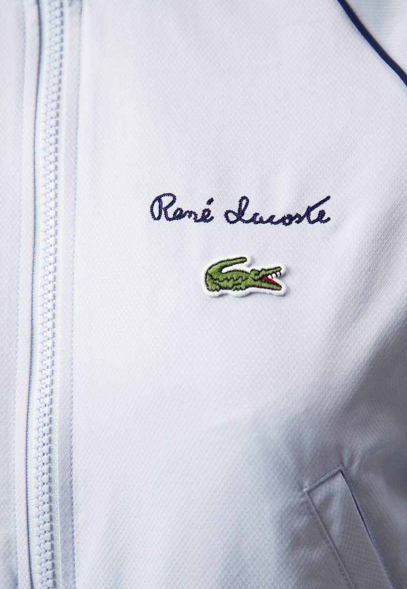 Hellblaues Reißverschlussjacke aus strukturiertem Stoff mit aufgestickter 'René Lacoste'-Signatur und grünem Krokodil-Logo auf der Brust.