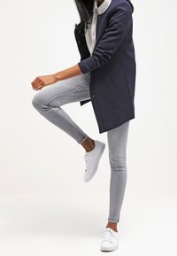 Marinblå kappa med avslappnad passform, i kombination med ljusgrå skinny jeans och vita sneakers. Kappan har knäppning och långa ärmar.