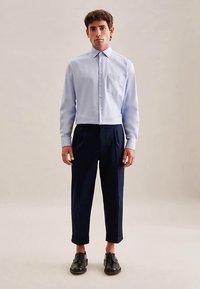 Camicia azzurro chiaro con bottoni e taschino sul petto, abbinata a pantaloni blu navy risvoltati alle caviglie e scarpe in pelle nera. Tessuto liscio.