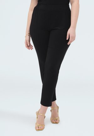 Person trägt schwarze körpernahe Hose und goldene Sandalen mit hohen Absätzen, steht mit gekreuzten Beinen vor einem einfarbigen weißen Hintergrund.