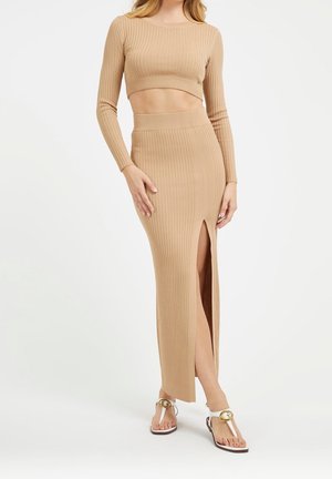 Frau trägt beigefarbenes, geripptes, langärmliges Crop-Top und passenden hochtaillierten Rock mit Frontschlitz, kombiniert mit weißen Sandalen mit goldener Schnalle.