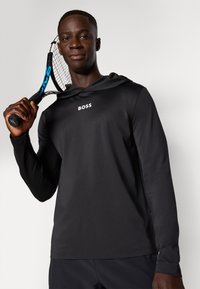 Musta pitkähihainen huppariurheilupaita, jossa on sileä pinta; rintataskussa valkoinen "BOSS"-logo; pidetään tennismailan kanssa.