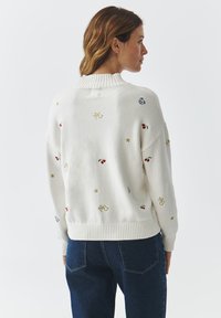 Witte gebreide trui met een geribde kraag en zoom, voorzien van kleurrijke geborduurde motieven zoals kersen, sterren en letters op een gladde textuur.