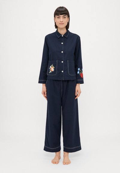 Conjunto de pijama de dos piezas en color azul marino que incluye una camisa con botones y bordado en los bolsillos, y pantalones de pierna ancha con costuras en contraste. Textura de tela suave.