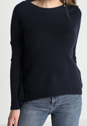 Femme portant un pull en tricot bleu marine foncé avec manches et ourlet côtelés, associé à un jean en denim bleu clair.