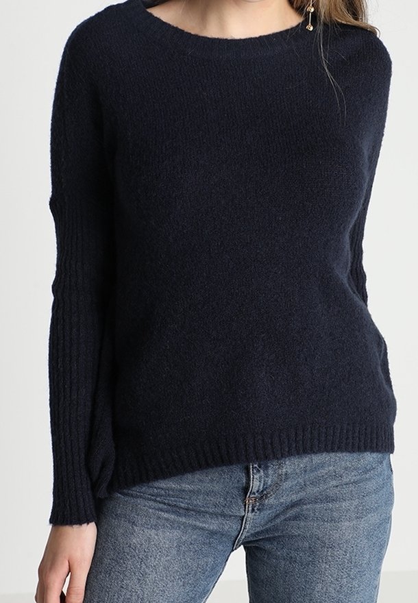 Femme portant un pull en tricot bleu marine foncé avec manches et ourlet côtelés, associé à un jean en denim bleu clair.