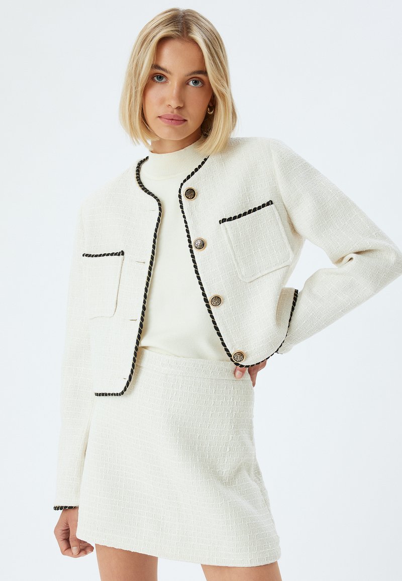 Giacca bianca strutturata con texture, arricchita da rifiniture e bottoni neri, abbinata a una gonna bianca con la stessa texture. Entrambi i capi hanno un design strutturato e sartoriale.