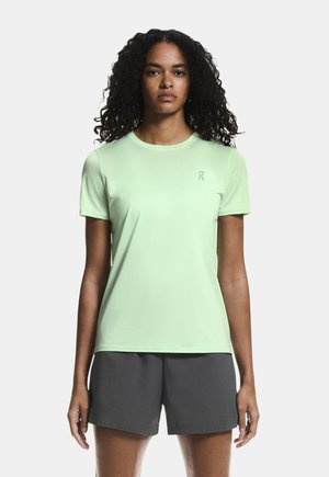 Woman with curly hair wearing a light green short-sleeve athletic shirt and dark gray shorts, standing against a plain background.