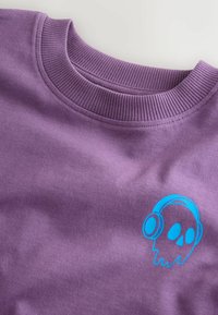 Violetinis džemperis su raštuota apykakle ir mėlyna grafika, vaizduojančia kaukolę su ausinėmis, kairiajame krūtinės pusėje. Lygi tekstūra.