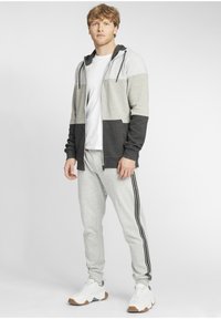 Grå och svart zip-up hoodie med en vit T-shirt under, kombinerat med ljusgrå träningsoverallsbyxor med svarta sidostråkar. Avslappnade sportskor.
