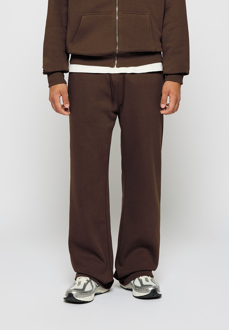 Brun bomullssweatshirt og matchende bukser, med glidelås foran, lommer og bred ben design, lagt over en hvit t-skjorte.