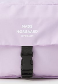 Lila tygväska med svart spänne och vit logotyp. Texturen är slät med en lätt lyster. Minimalistisk design.