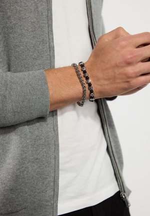 Zilveren schakelsymbool armband en zwarte kralenarmband om de pols. Grijze, met rits gesloten cardigan met geribbelde manchetten. Witte T-shirt eronder.