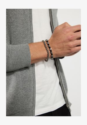 Bracelete de corrente prateada e bracelete de contas pretas no pulso. Cardigã cinza com fecho éclair e punhos em canelado. Camiseta branca por baixo.