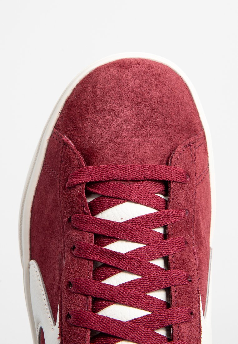 Sneaker in suede bordeaux con dettagli in pelle bianca, caratterizzata da lacci piatti e una punta tonda. Texture liscia e dettagli colorati a contrasto.