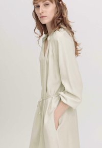 Hellgrünes Wickelkleid mit Dreiviertelarm, V-Ausschnitt und gebundenem Taillengürtel. Der Stoff wirkt weich mit einer glatten Textur und seitlichen Taschen.