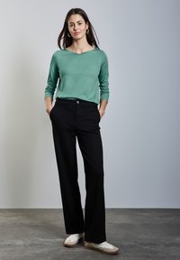 Vihreä pitkähihaine paita, jossa on leveä kaula-aukko, mustat muotoonommellut housut, joissa on etutaskut, sekä valkoiset lenkkarit, joissa on vaaleanruskeita yksityiskohtia.