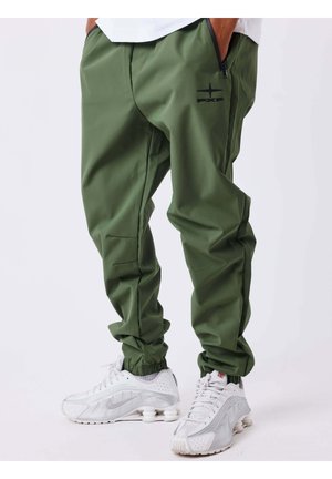 Personne portant un pantalon de jogging vert avec des poignets élastiques et un logo noir, associé à des baskets blanches Nike avec un amorti à air visible.