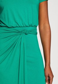 Partie médiane d'une personne portant une robe verte avec un détail de tissu noué à la taille et un bras découvert du côté droit.