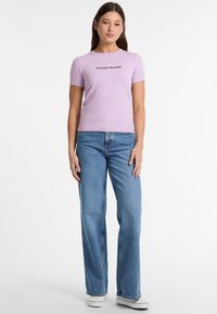 Šviesiai violetinė trumpa rankovė t-marškiniai su "GUESS JEANS" logotipu, derinami su mėlynomis platėjimo džinsais ir baltais sportbačiais, ant neutralios fono.