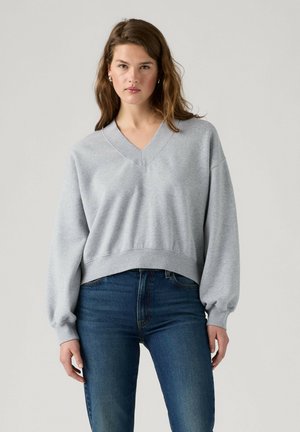 Ung kvinde med langt brunt hår iført en lys grå V-hals trøje og blå jeans, stående mod en ensfarvet baggrund.