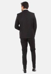 Completo nero con uno spacco, vestibilità su misura. Texture liscia, leggero luccichio. I pantaloni e le scarpe nere completano l'aspetto formale.