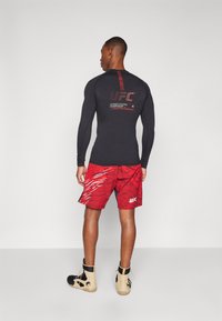 Chemise noire à manches longues avec le logo UFC et des accents rouges, associée à un short rouge ornée d'un motif graphique et d'un logo UFC, portée avec des chaussures.