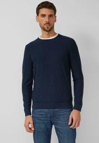 Man met kort bruin haar en getrimde baard, gekleed in een marineblauwe trui over een wit overhemd en een blauwe spijkerbroek, staand tegen een effen grijze achtergrond.