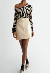 Top nero e beige con motivo, scollatura a barca e maniche lunghe, abbinato a una minigonna beige in ecopelle con cintura larga. Scarpe con dettagli dorati.