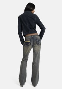 Γυναίκα που φορά μαύρο τζιν τζάκετ και γκρι τζιν παντελόνι με φουντωτά μπατζάκια, διακοσμημένο με αλυσίδες στις πίσω τσέπες, στέκεται μπροστά σε απλό λευκό φόντο.