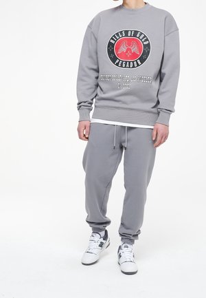 Sudadera gris con un gráfico redondo que presenta alas y texto, combinada con unos joggers grises a juego y zapatillas blancas. Material de suave mezcla de algodón.