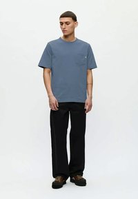 Νεαρός άνδρας με μπλε κοντομάνικο μπλουζάκι με τσέπη, μαύρο παντελόνι σε φαρδιά γραμμή και καφέ δερμάτινα παπούτσια με κορδόνια, που στέκεται μπροστά σε λευκό φόντο.