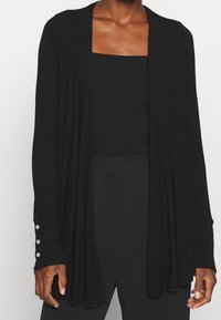 Kobieta w czarnym kardiganie zapinanym na przód, nosząca czarny top z prostokątnym dekoltem oraz czarne spodnie z wysokim stanem, stoi z rękami w luźnej pozycji.