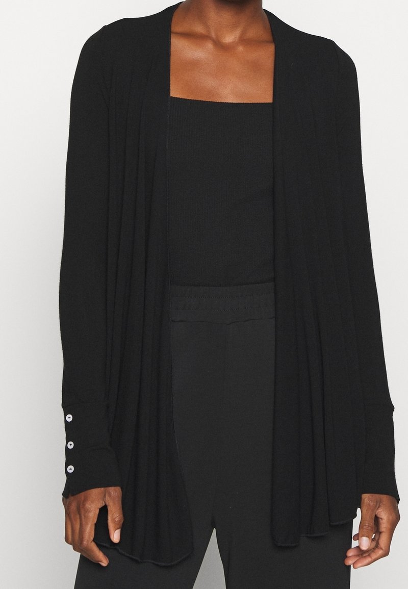 Kobieta w czarnym kardiganie zapinanym na przód, nosząca czarny top z prostokątnym dekoltem oraz czarne spodnie z wysokim stanem, stoi z rękami w luźnej pozycji.