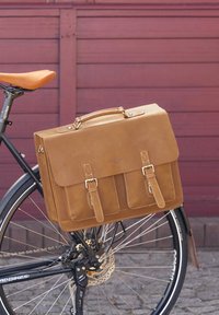 Hnedá kožená messenger taška s dvojitým zapínaním na pracky, šitím a hornou rúčkou, umiestnená na čiernom bicykli proti červenému pozadiu.