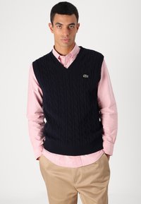 Σκούρο μπλε πλεχτό γιλέκο με πλέξη καλωδίωσης, σχέδιο v-neck και ραμμένο στρίφωμα, φορεμένο πάνω από ένα ανοιχτό ροζ πουκάμισο με κουμπιά και μπεζ παντελόνι. Λεπτομέρεια πράσινου λογότυπου.