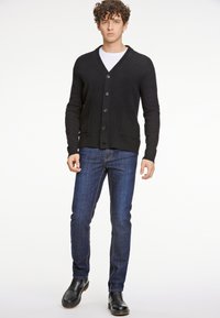 Svart stickad kofta med V-hals, knappt stängning och framfickor, ihopparad med blå jeans och svarta ankelboots.