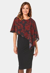 Rode cape met bloemenpatroon over een zwarte getailleerde jurk, met een vloeiend ontwerp, een doorschijnende textuur en versierde randen.