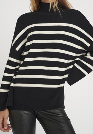 Pull en noir et blanc à rayures avec col roulé, coupe oversize, texture côtelée et manches larges, associé à un pantalon en cuir synthétique noir.